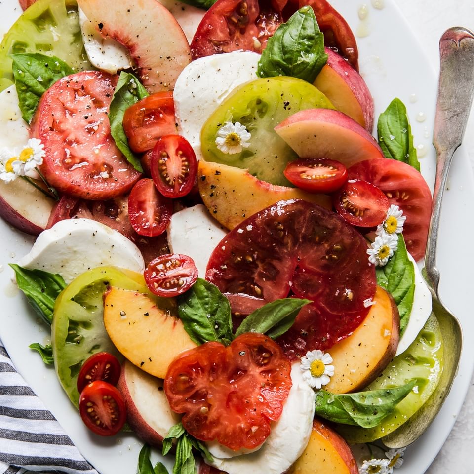 summer peach and tomato salad with fresh mozzarella, basil and olive oil on a large white platter.
