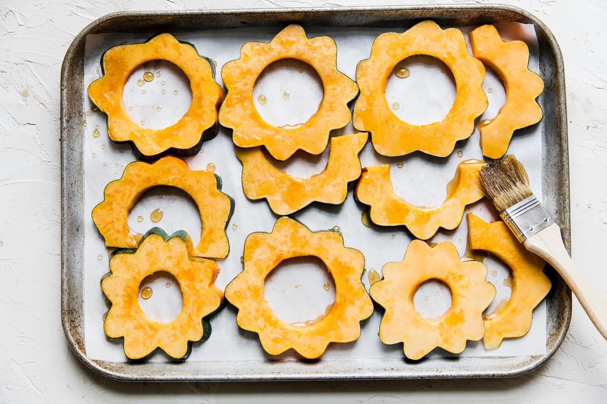 sliced acorn squash on a baking sheet being brushed with maple syrup