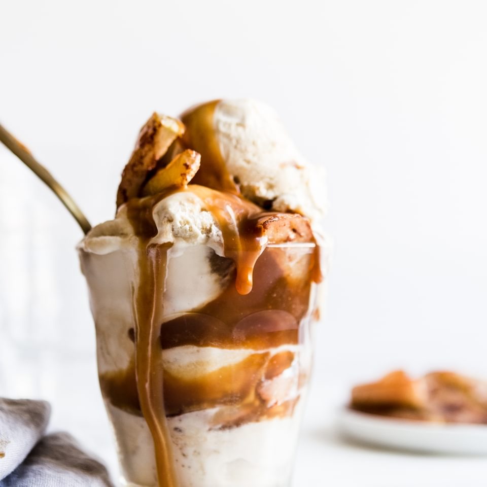 A vanilla ice cream and roasted apple sundae in a cup with a spoon and salted caramel dripping over the side.