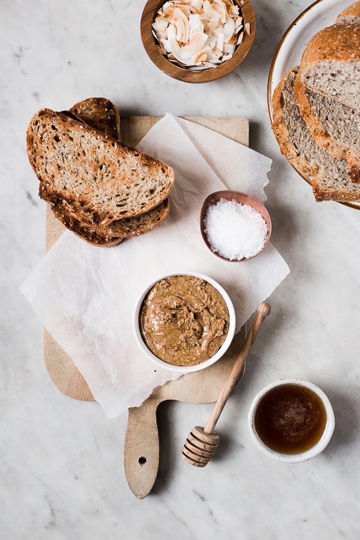 Slices of bread on a cutting board next to a bowl of almond butter, honey , toasted coconut flakes and flakey salt.