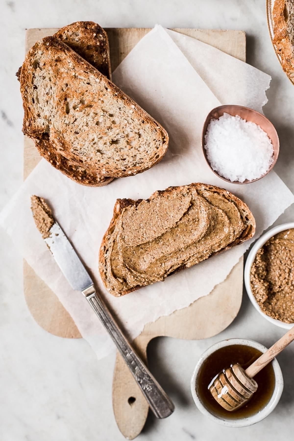 A slice of toasted bread spread with almond butter. Next to a bowl of honey, flakey salt and a knife.