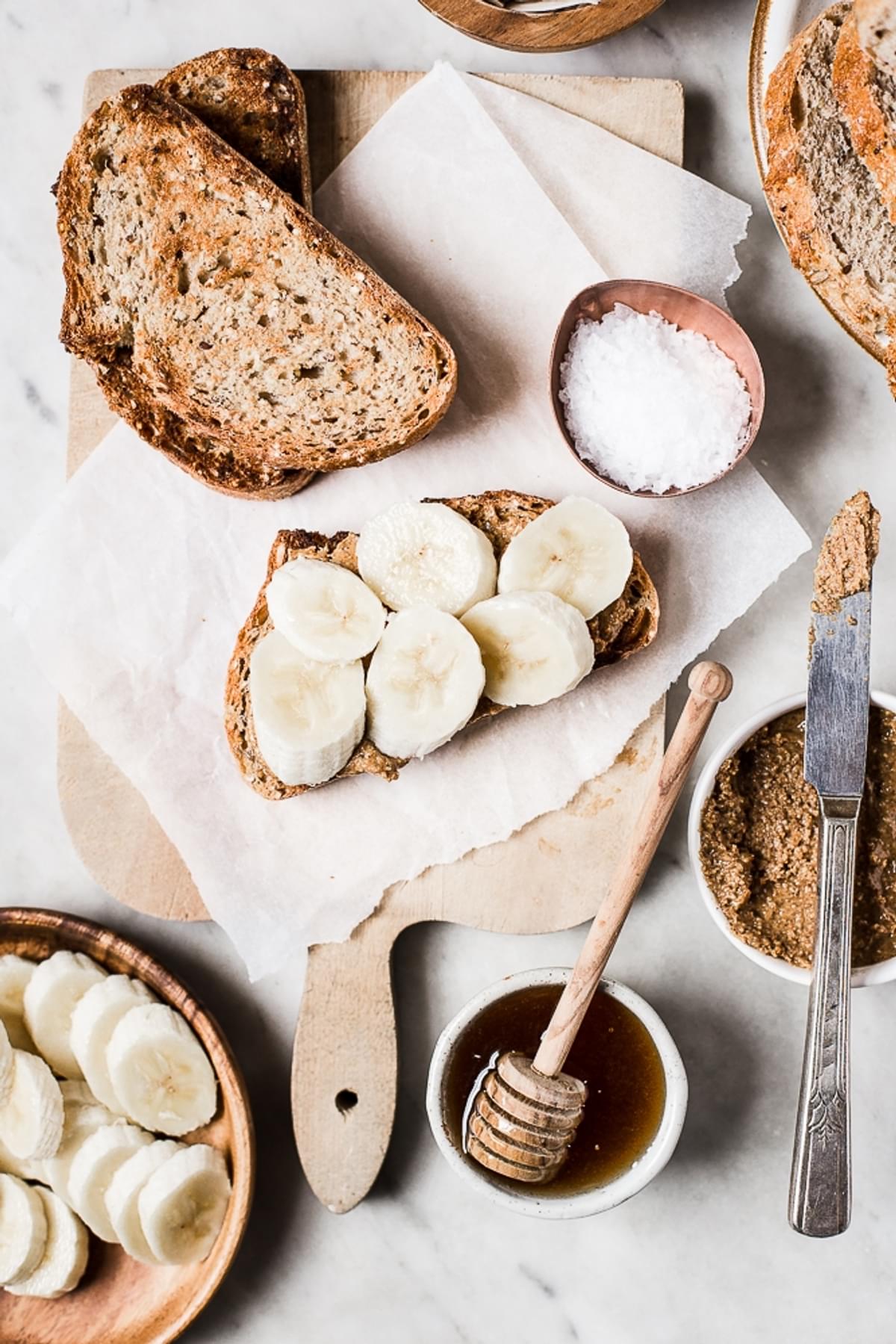 Almond Butter Toast with Banana slices on a cutting board next to a bowl of honey and a bowl of flakey salt.