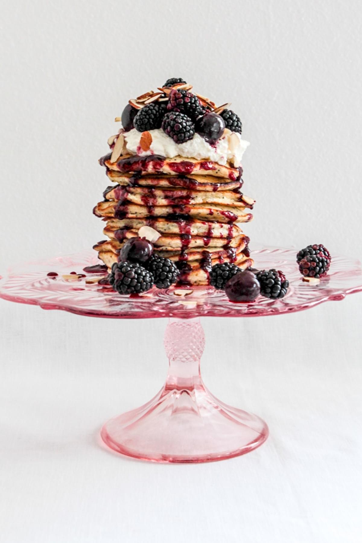 a stack of almond poppy seed pancakes topped with whipped cream, fresh berries and maple syrup on a pink cake plate