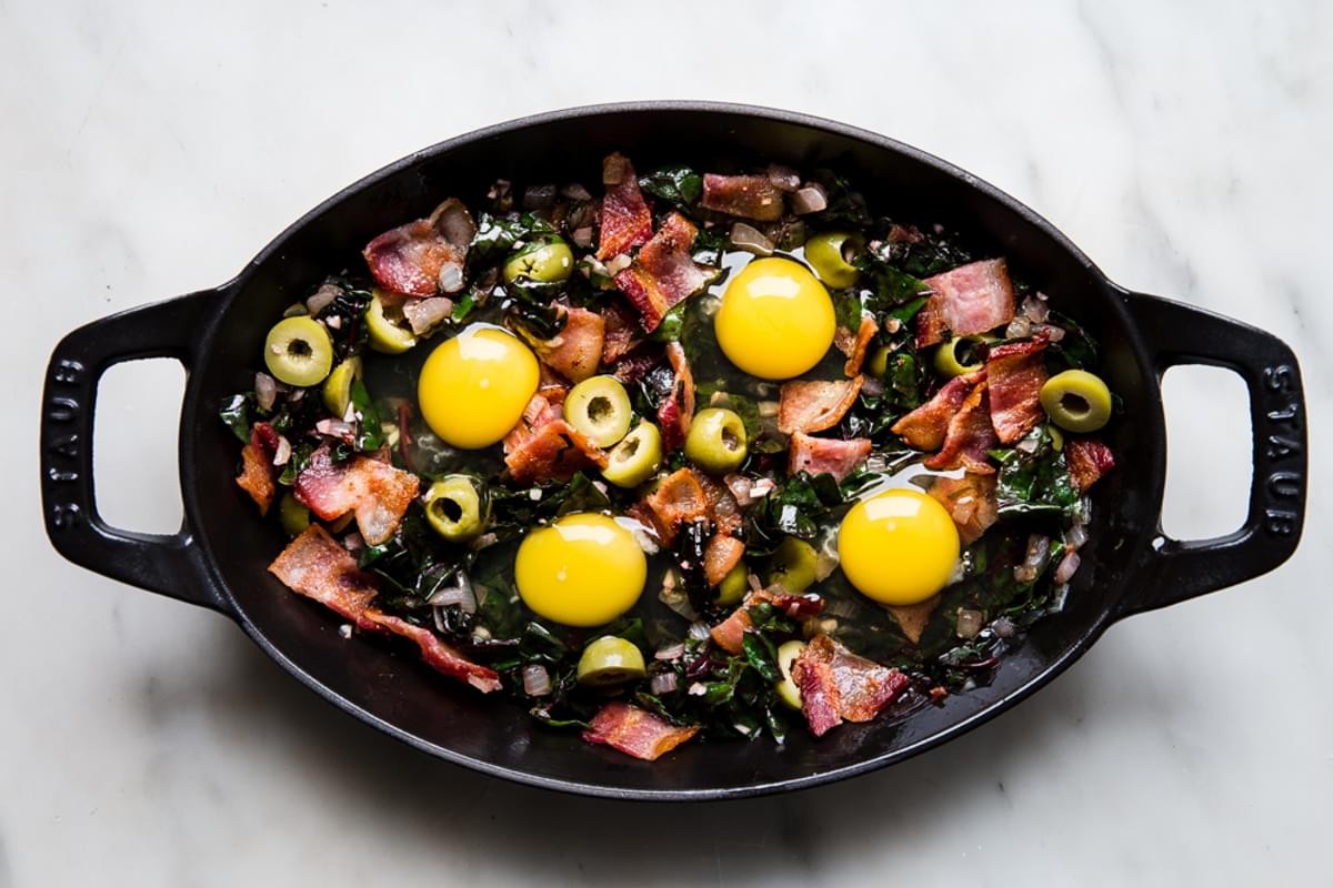 raw eggs cracked into sautéed onions and garlic in a pan with swiss chard, bacon and green olives