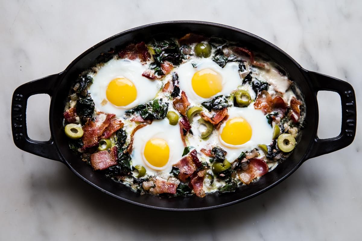 eggs baked into sautéed onions and garlic in a pan with swiss chard, bacon and green olives