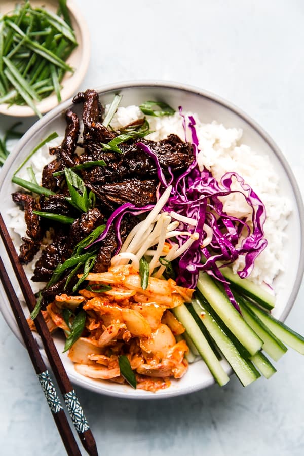 a bowl of cooked white rice topped with beef bulgogi, kimchi, bean sprouts, red cabbage and cucumbers