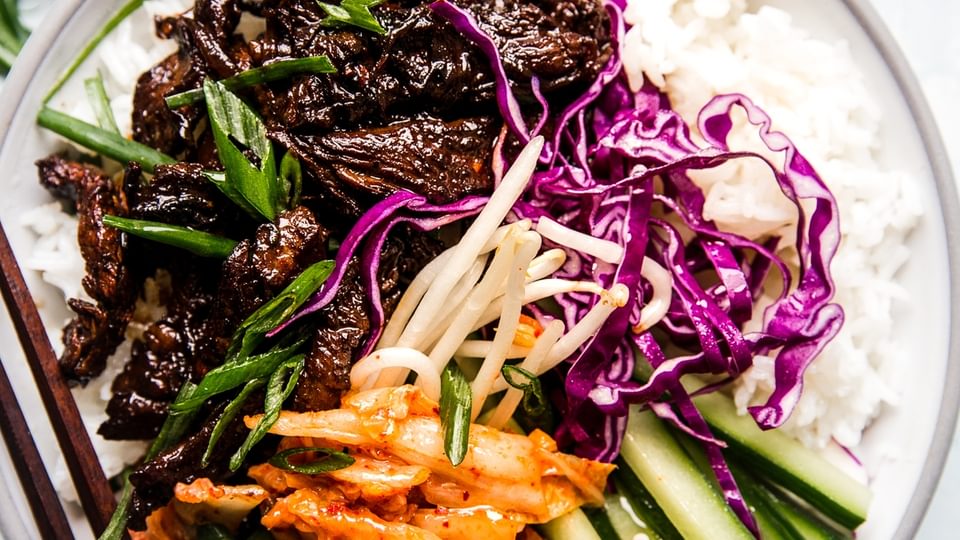a bowl of cooked white rice topped with beef bulgogi, kimchi, bean sprouts, red cabbage and cucumbers