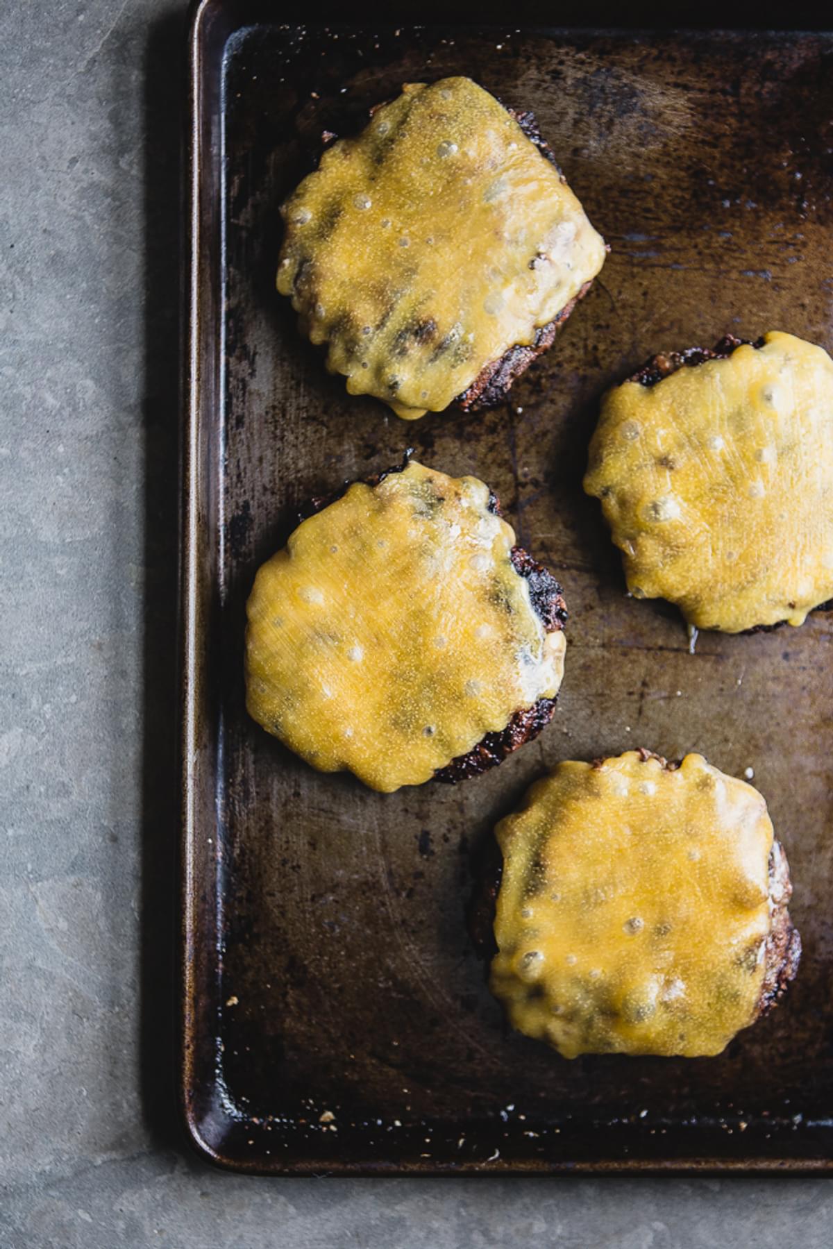 4 cooked beef patties on a baking sheet topped with melted cheddar cheese