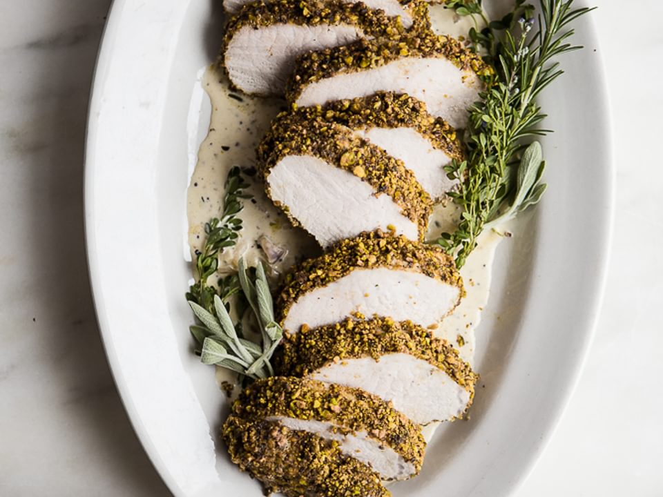 Pistachio Crusted Pork Tenderloin with White Wine Dijon Sauce on a plater with herbs and extra sauce in a dish