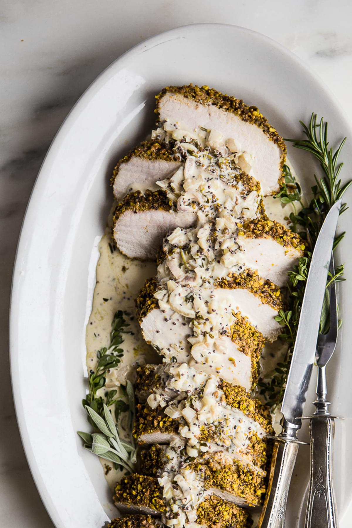 Pistachio Crusted Pork Tenderloin with White Wine Dijon Sauce on a plater with a knife and fresh herbs