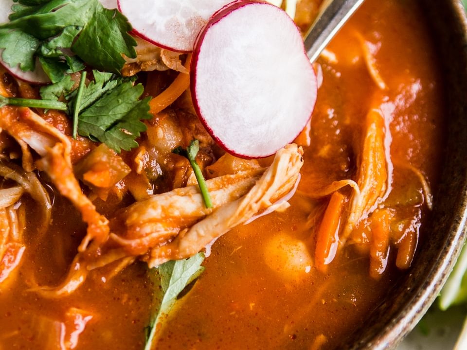 turkey pozole rojo in brown bowl topped with cilantro, radish slices and shredded cabbage