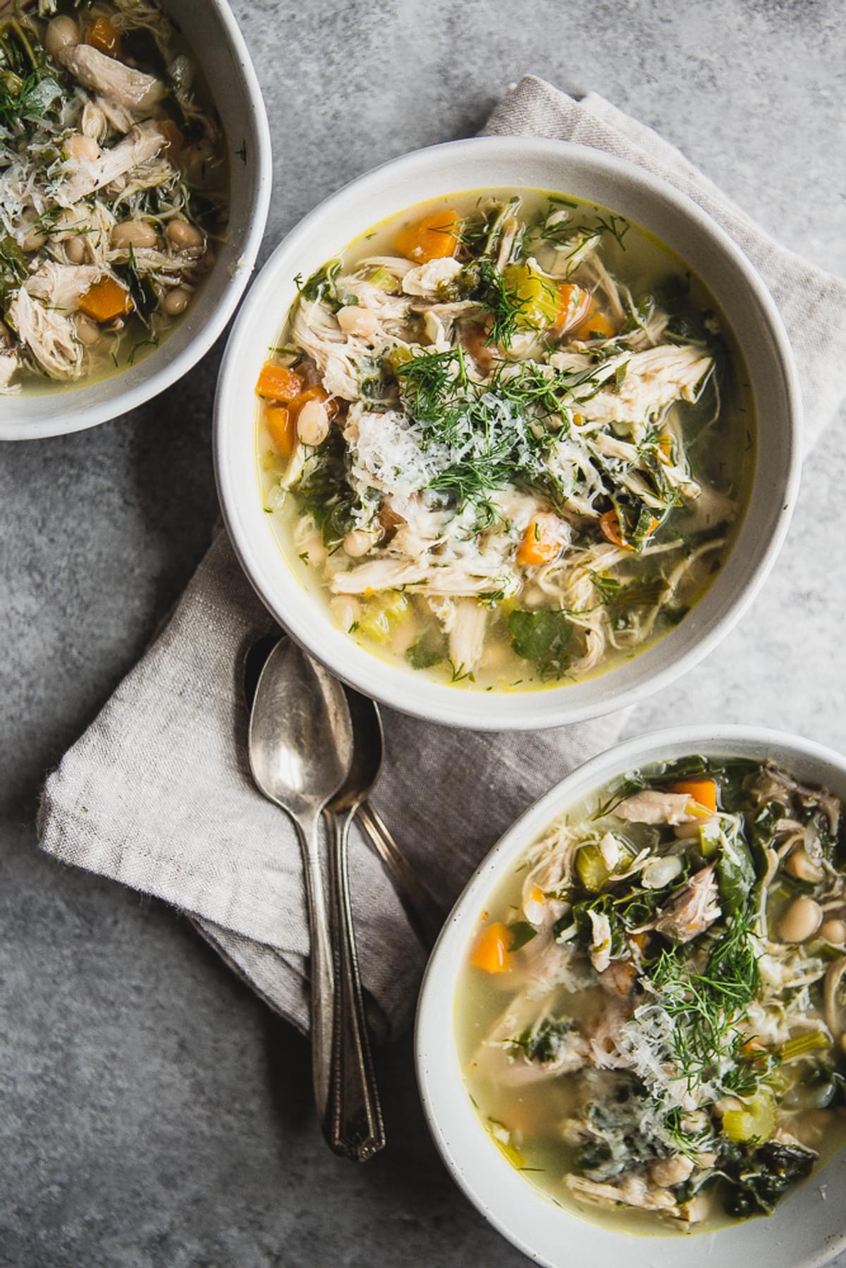 three bowls of white bean chicken soup with spoons and a linen