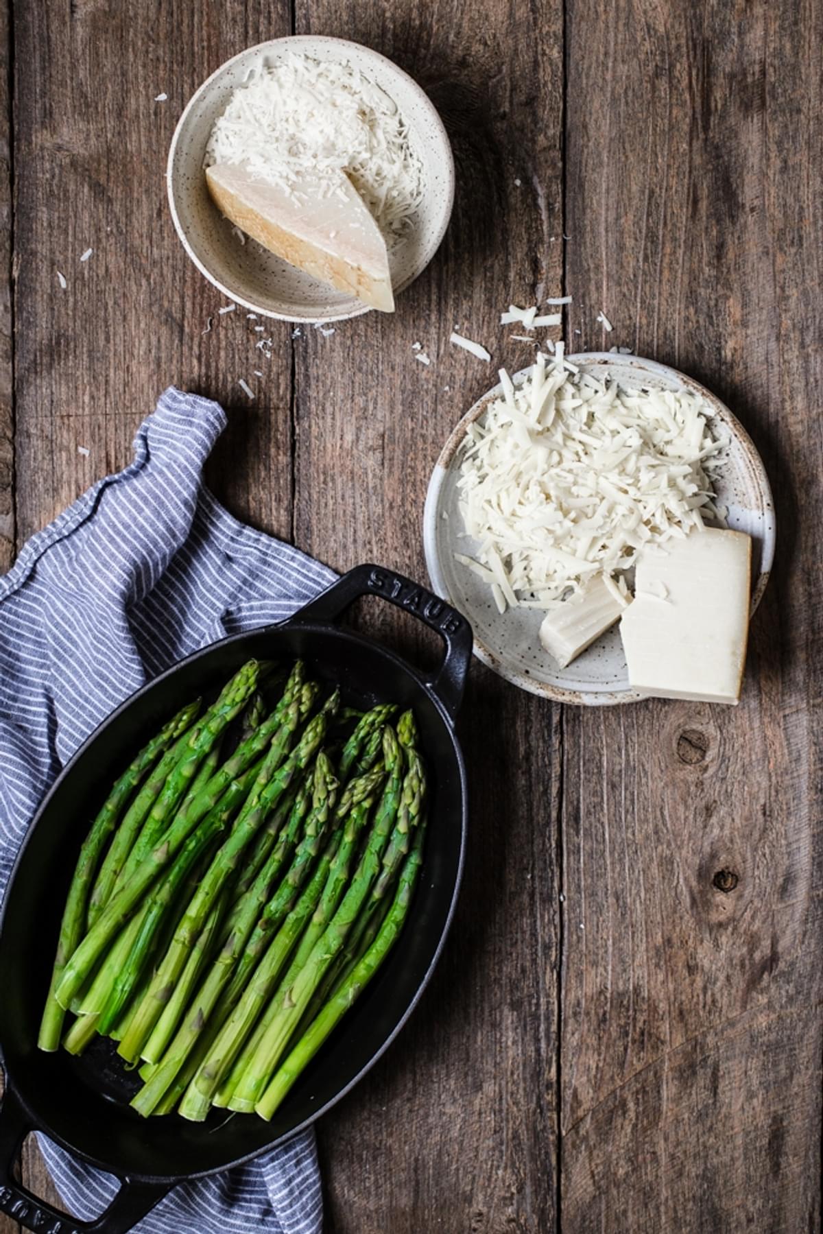 blanched asparagus in a cast iron baking dish next to a plate of grated gruyère cheese