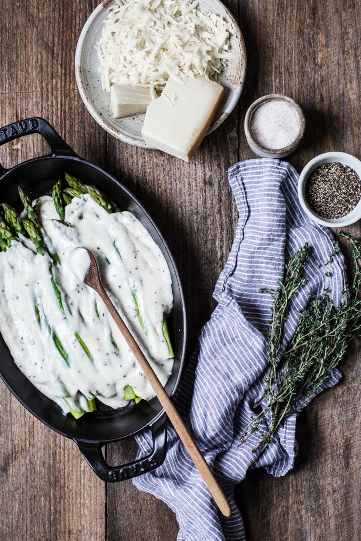 blanched asparagus in a small cast iron baking dish topped with a creamy béchamel sauce next to a plate of shredded Gruyère