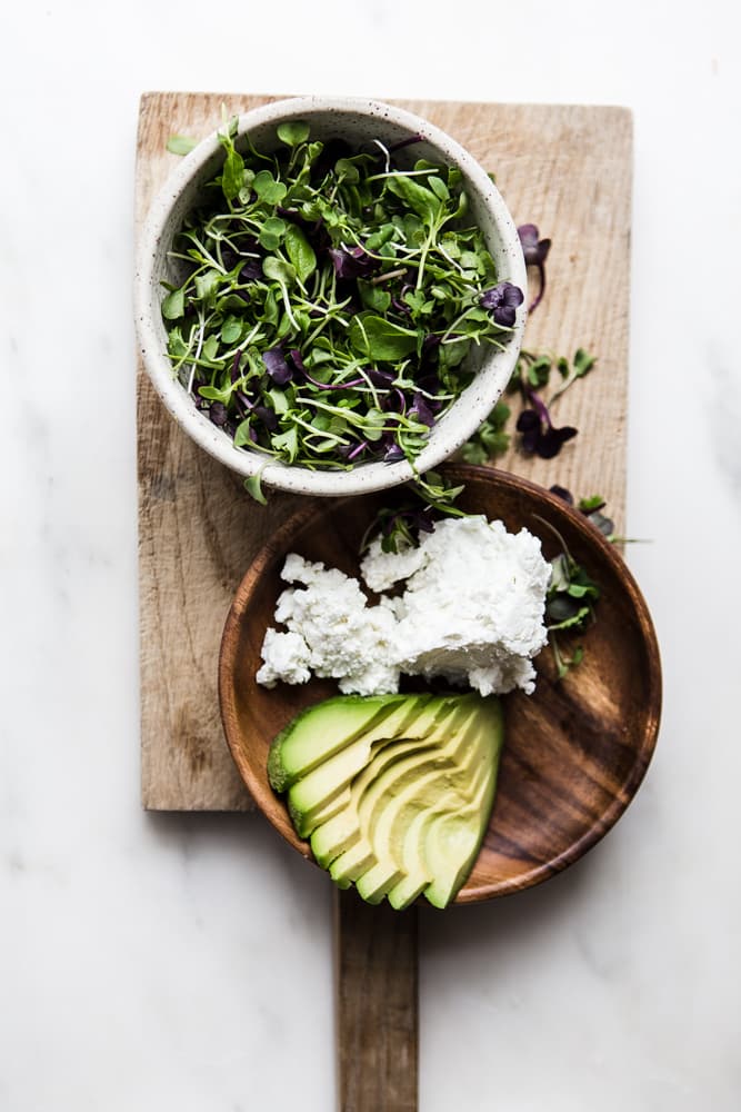 a plate of avocado and goat cheese and a bowl of micro greens on a cutting board