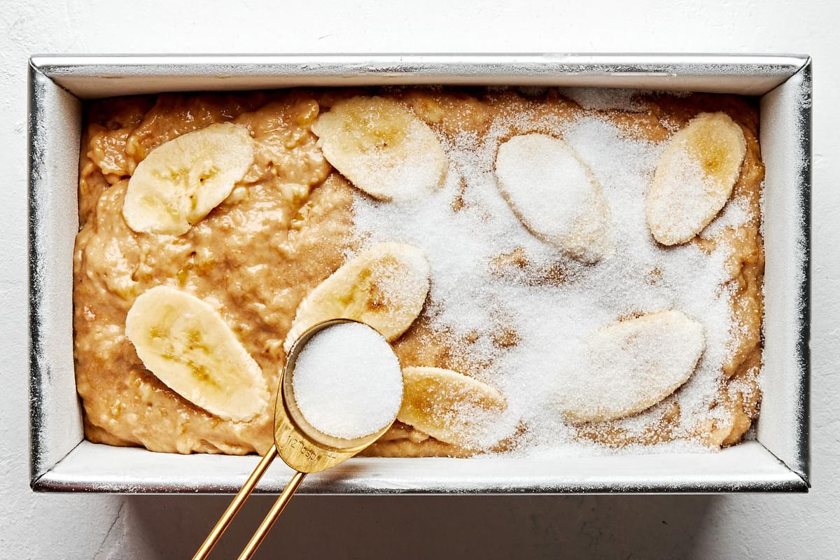 banana bread dough in a prepared loaf pan being topped with thinly sliced bananas and sprinkled with granulated sugar