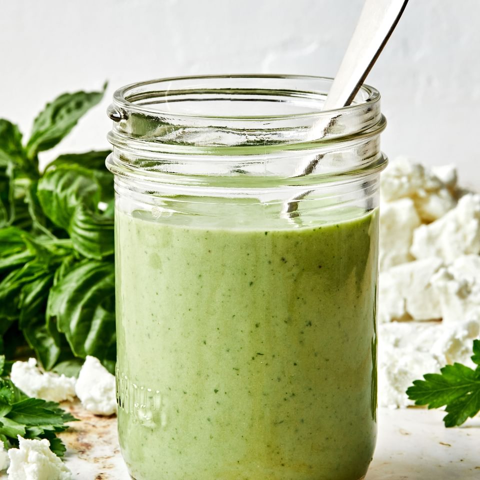 Homemade green goddess dressing in a glass jar with a spoon surrounded by fresh basil on the counter