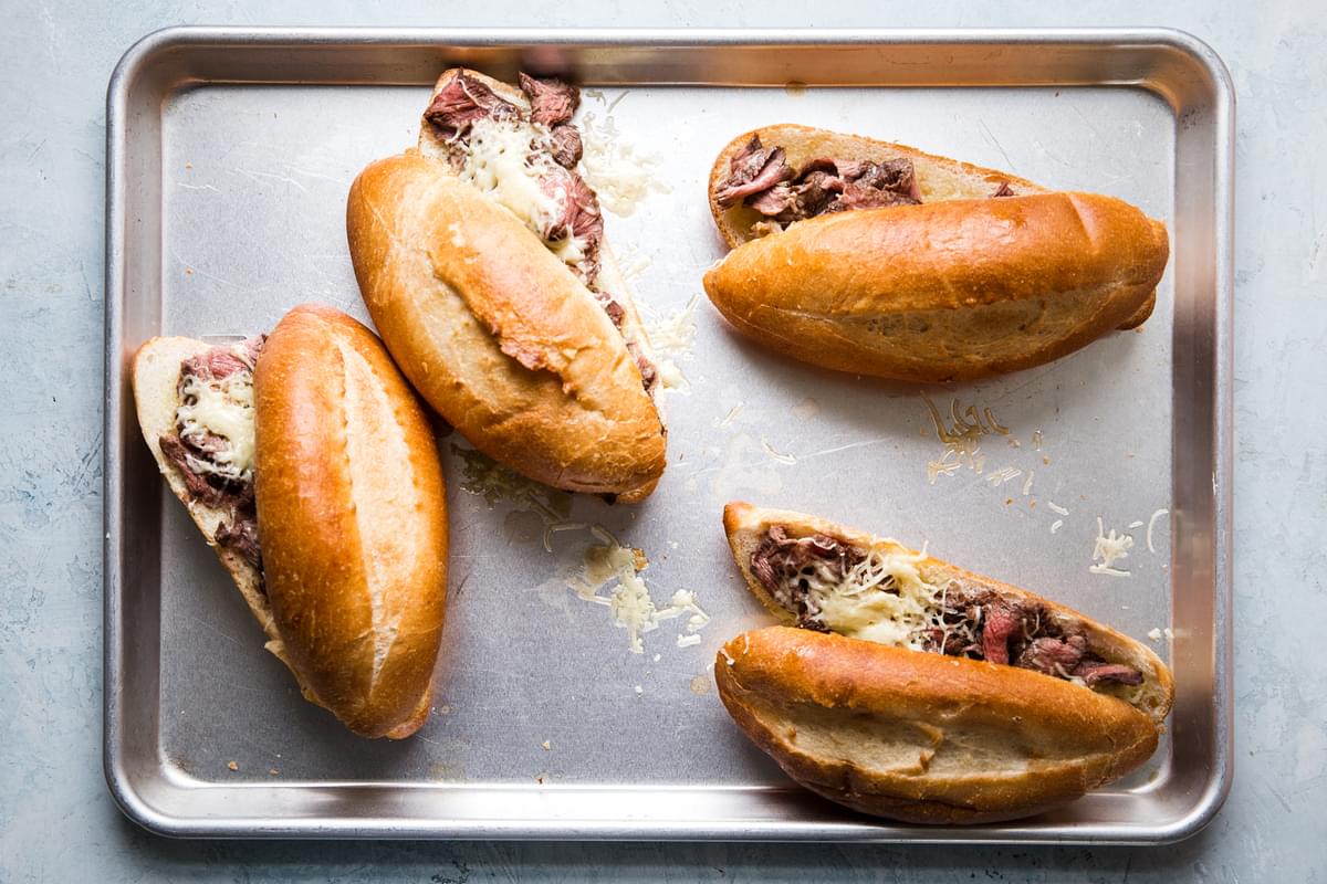 four homemade french dip sandwiches on a baking sheet with thinly sliced steak, garlic butter and gruyere cheese