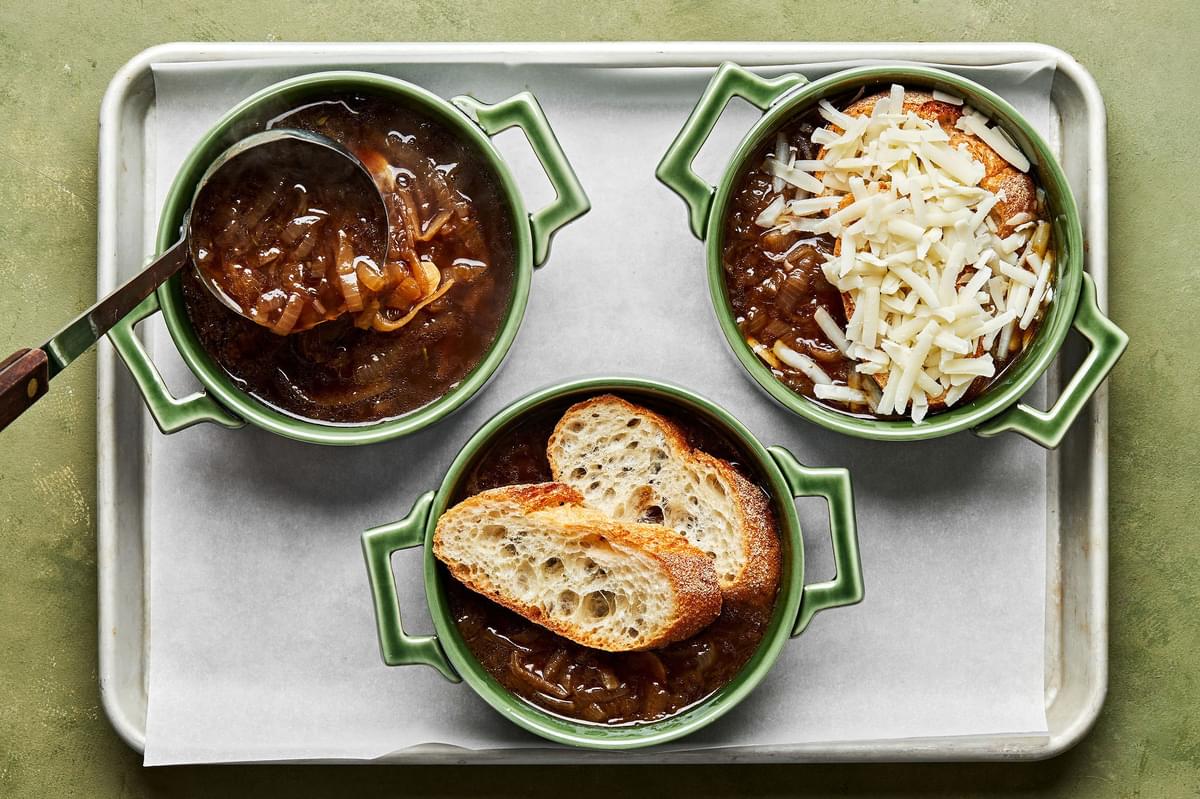 3 small oven proof bowls of french onion soup being topped with sliced baguette and Gruyère cheese