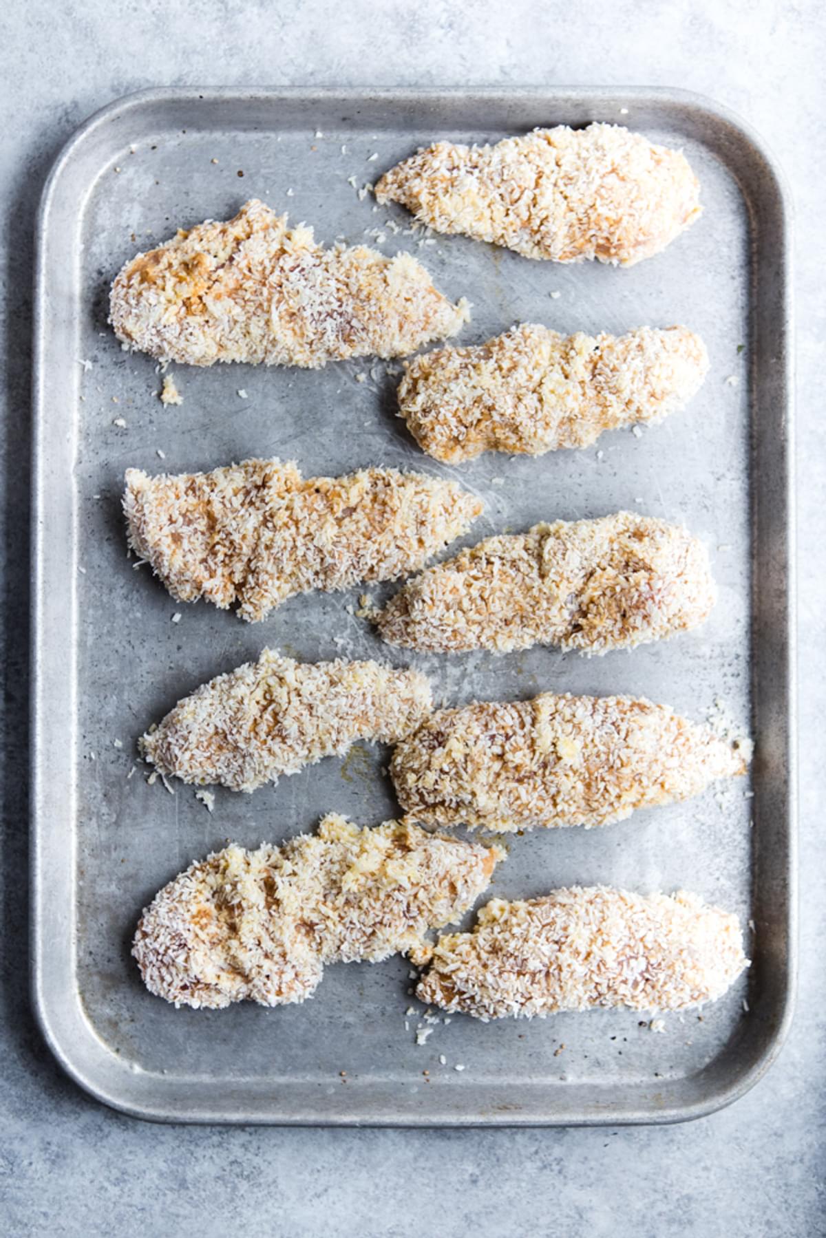 raw chicken tenders wit almond flour and coconut on a baking sheet