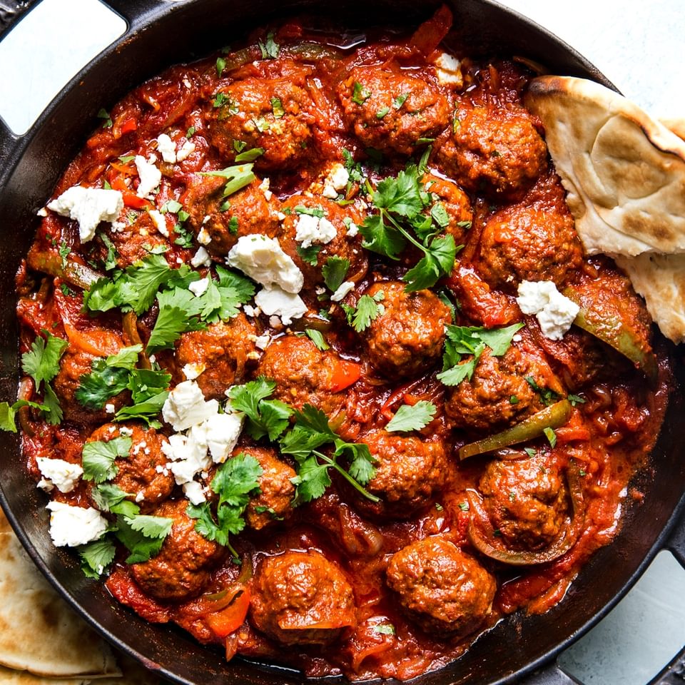 spiced meatballs simmered in a shakshuka sauce in a skillet served with feta, parsley and pita bread