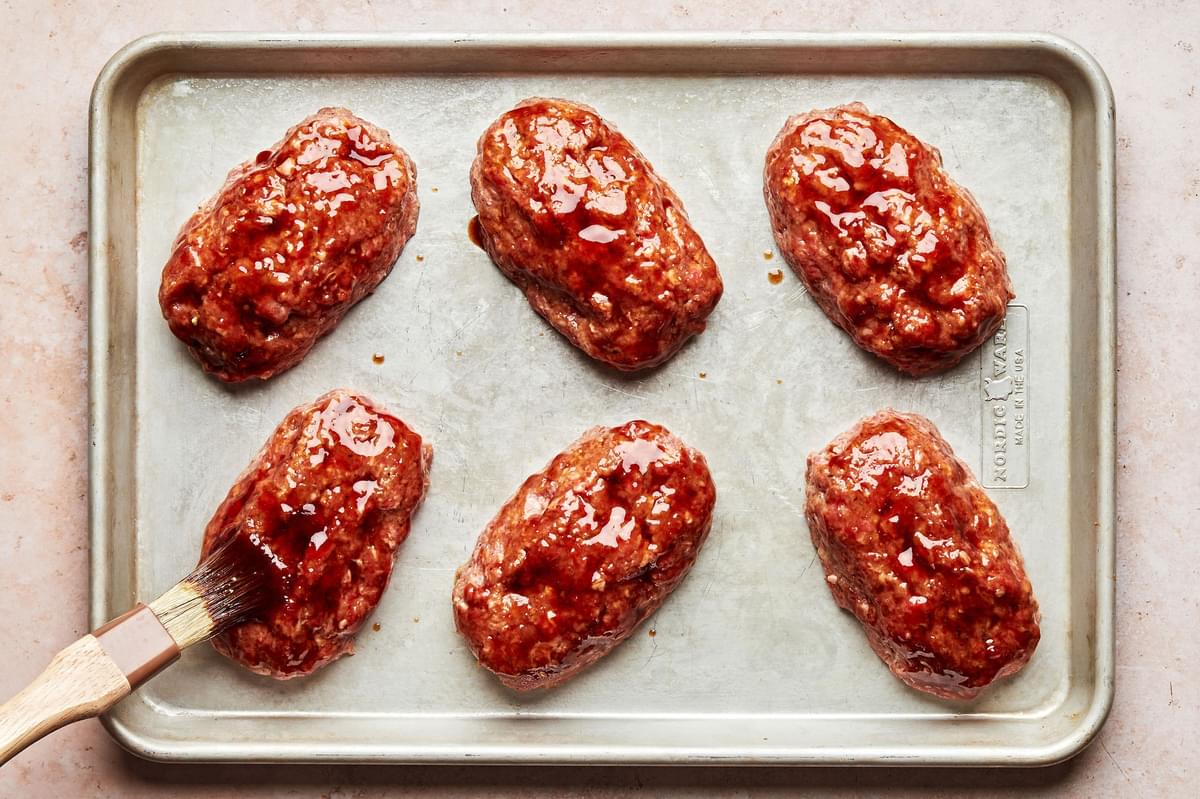 6 mini meatloaves on a baking sheet being brushed with a brown sugar sauce on top before baking