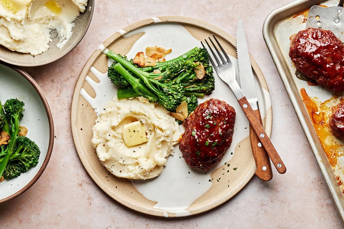 a plate with mini meatloaf served with mashed potatoes and broccolini  surrounded by serving bowls of potatoes and meatloaf