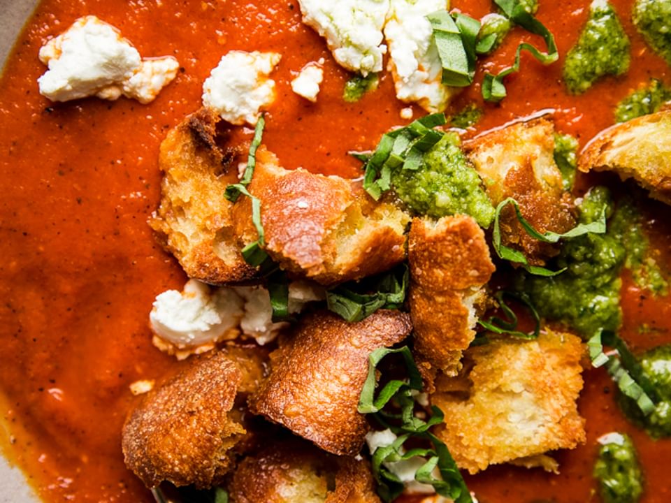 Roasted red pepper soup in a bowl with croutons, goat cheese and pesto with a spoon