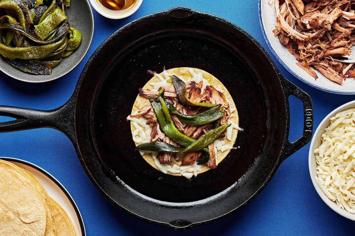 a corn tortilla topped with carnitas, roasted poblanos and mozzarella cheese being cooked in a skillet