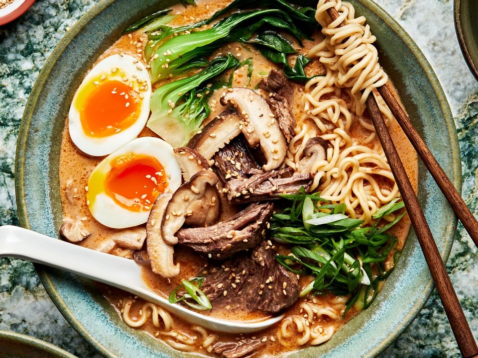 a bowl of slow cooker curry beef ramen topped with soft boiled eggs with a spoon and chopsticks