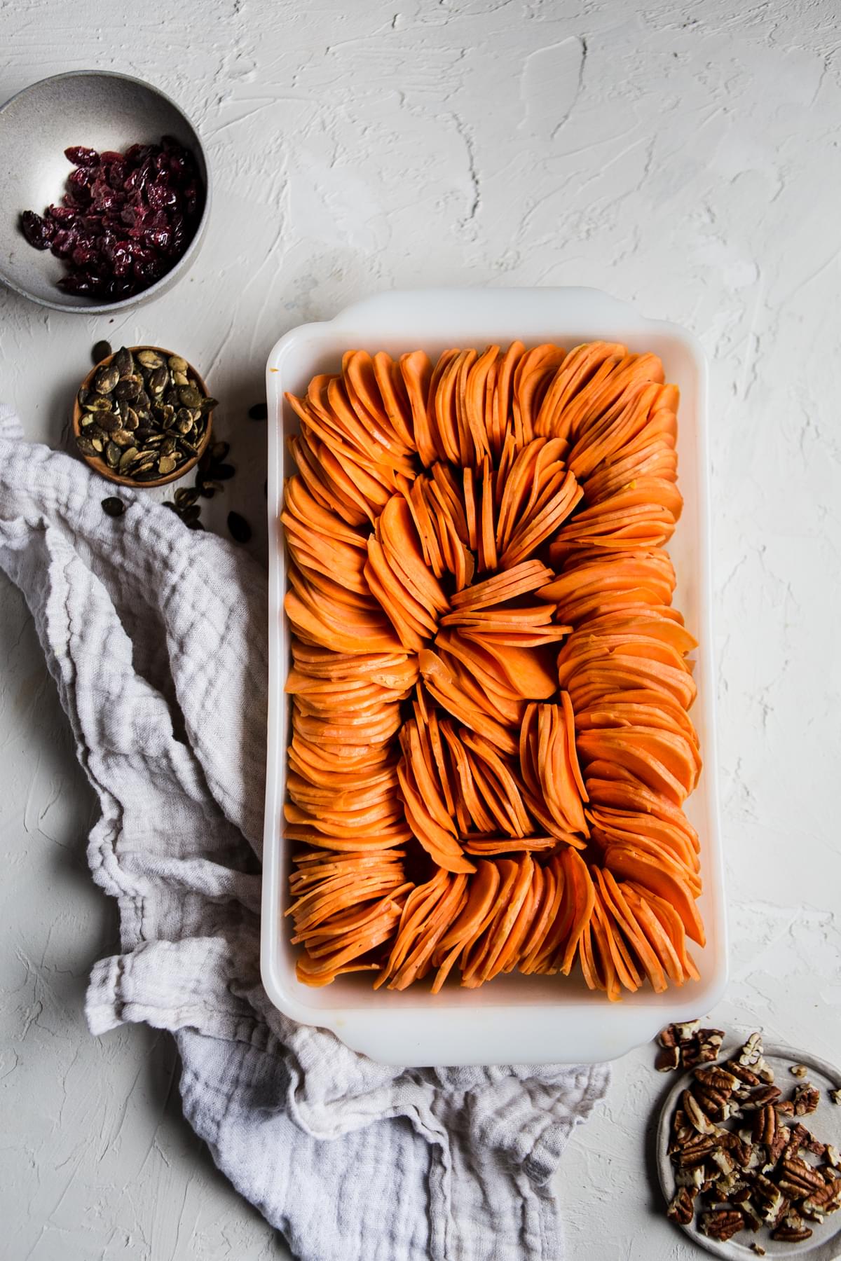 Sweet potato slices arranged in a baking dish next to bowls of dried cranberries, pumpkin seeds and pecans