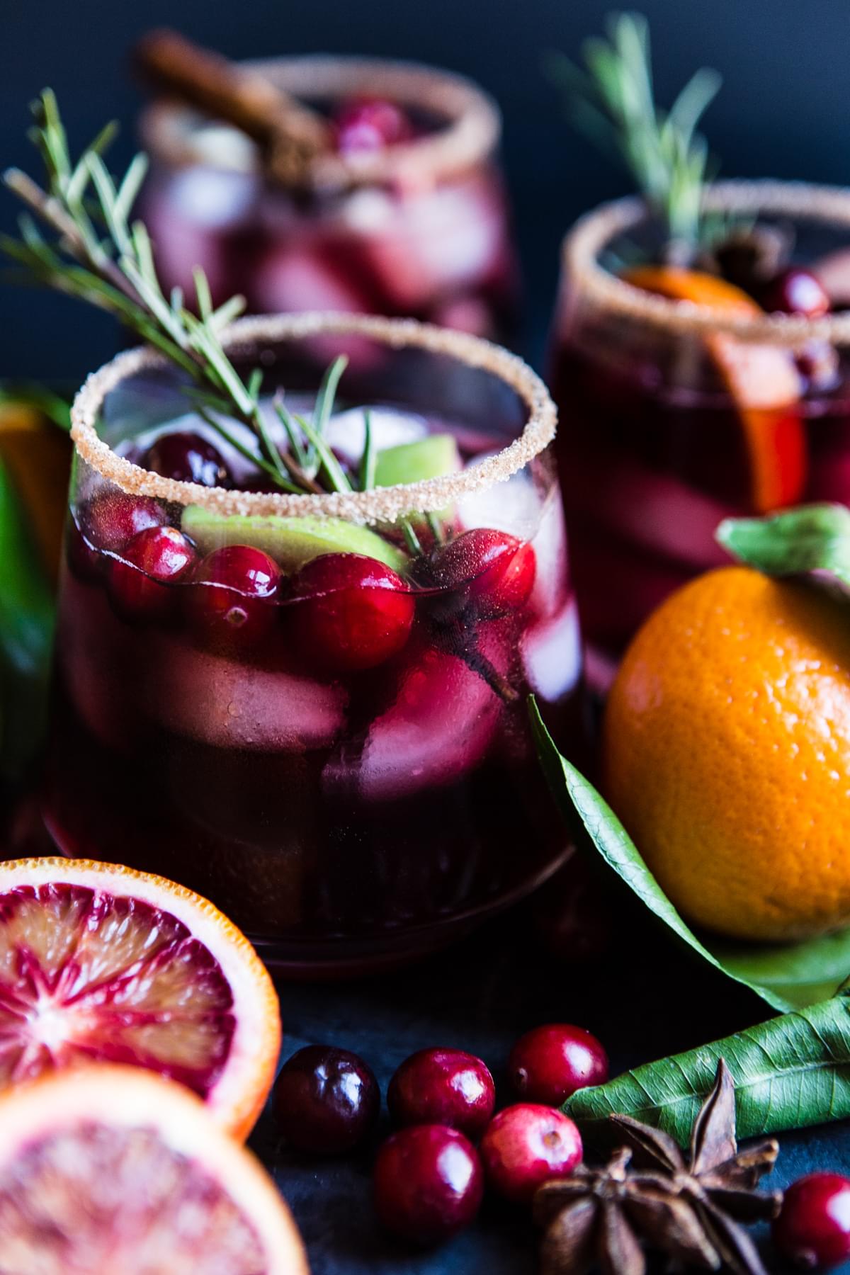 3 glasses filled with red wine sangria made with cranberries, apples, oranges, cinnamon and brandy garnished with rosemary