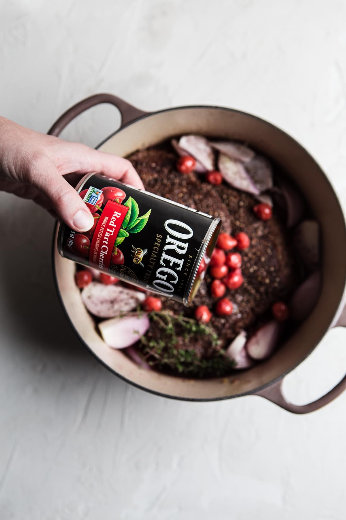 a hand pouring red berries from a can into a pot with a beef roast and shallots in it