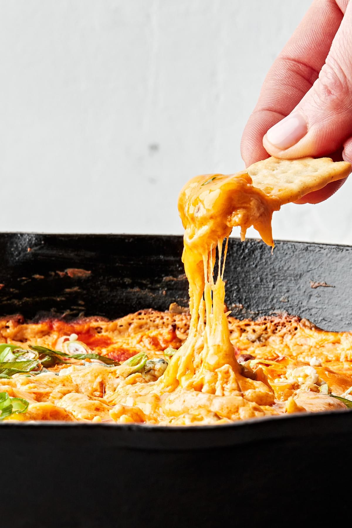 hand dipping chicken flavored cracker into a cast iron skillet full of homemade buffalo chicken dip