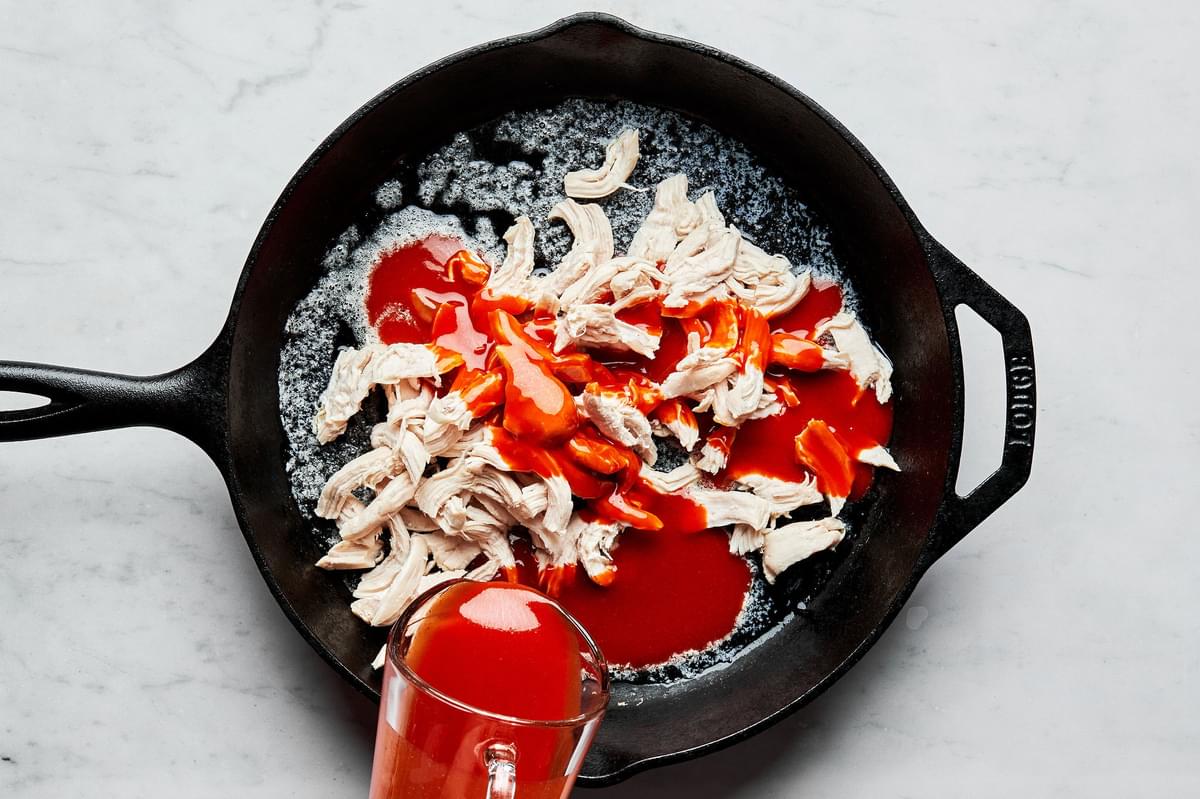 buffalo sauce being poured into a cast iron skillet with melted butter and cooked shredded chicken for buffalo chicken dip