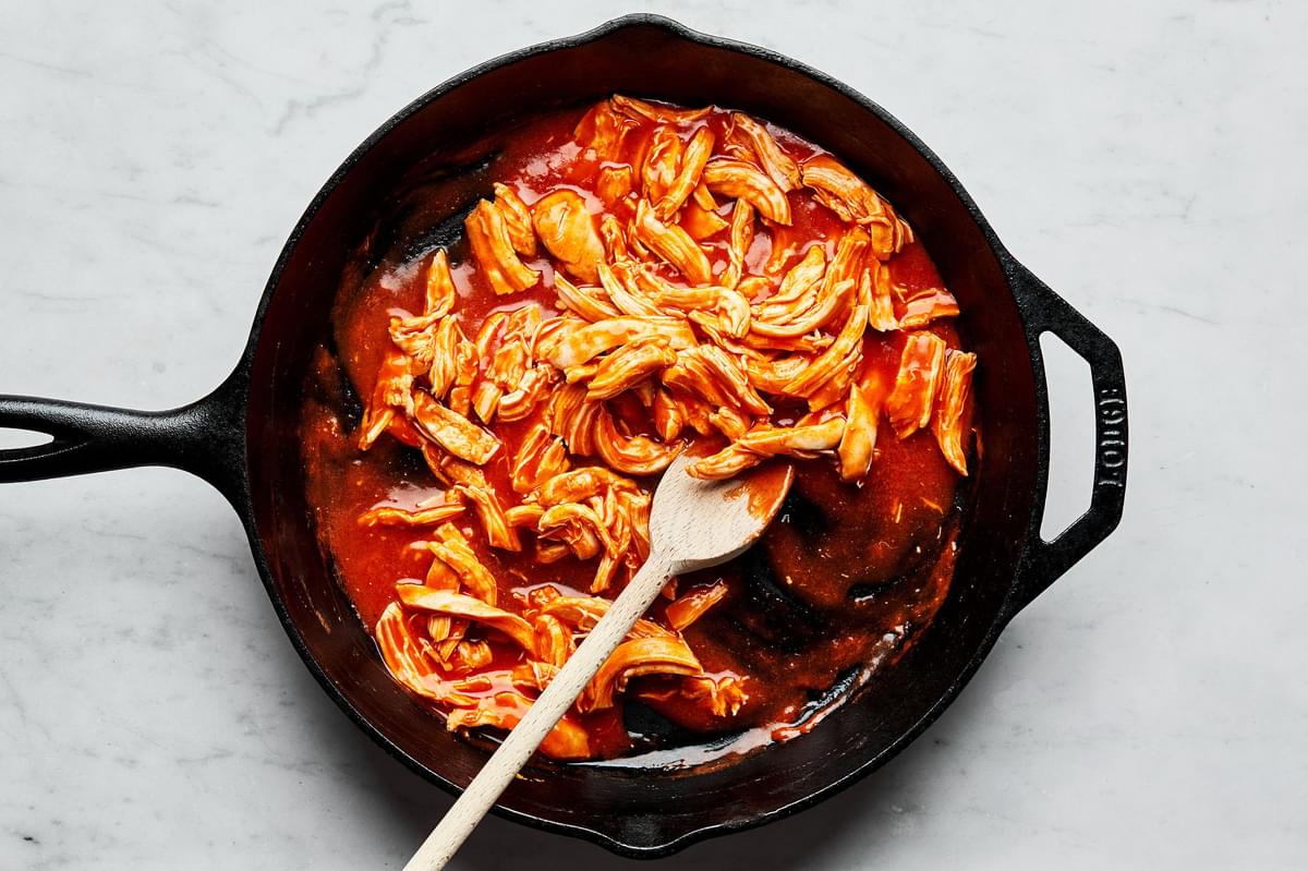 buffalo sauce, cooked shredded chicken and melted butter being stirred together with a wooden spoon in a cast iron skillet