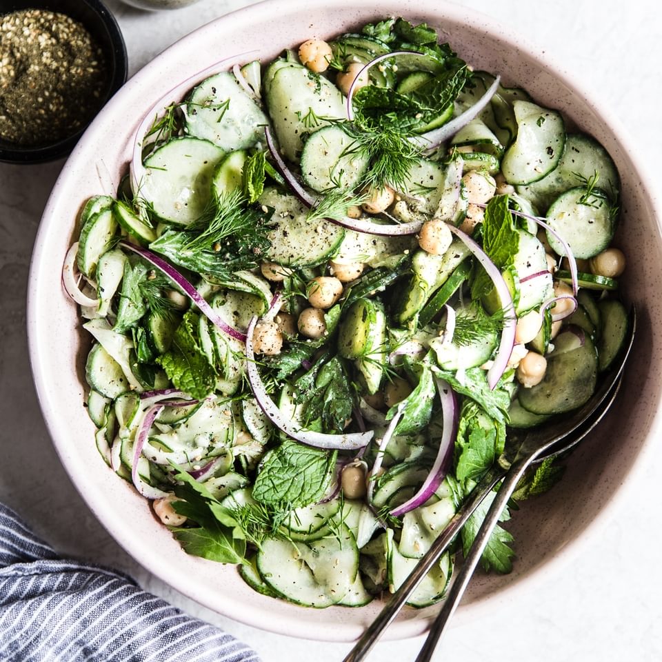 cucumber salad with  chickpeas, onion, parsley, mint, dill and a yogurt dressing in a bowl with serving spoons