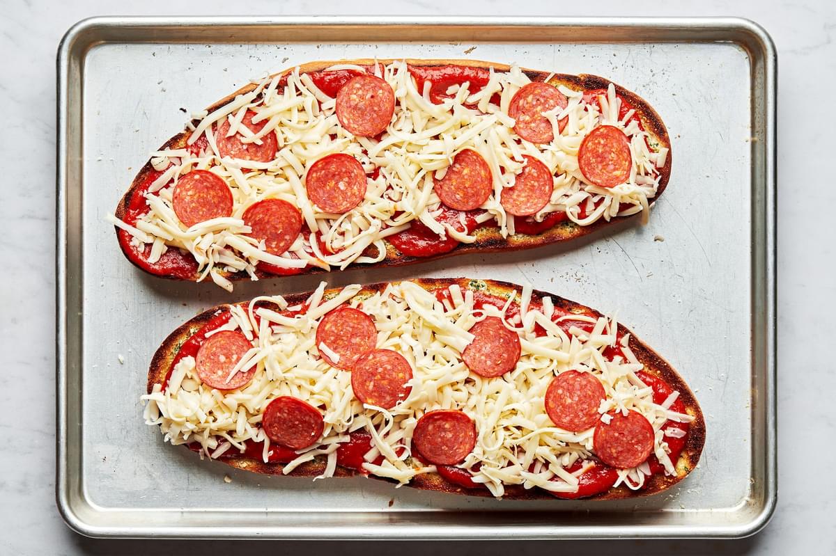 french bread sliced in half lengthwise topped with butter, spices, pizza sauce, mozzarella and pepperoni on a baking sheet