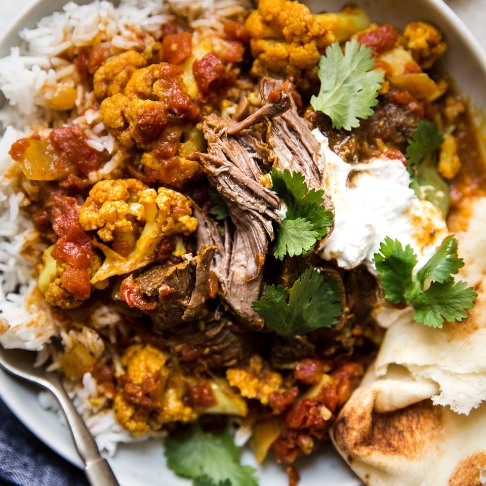 A bowl of Indian Style Lamb Curry With Cauliflower over white rice and served with yogurt and cilantro and naan