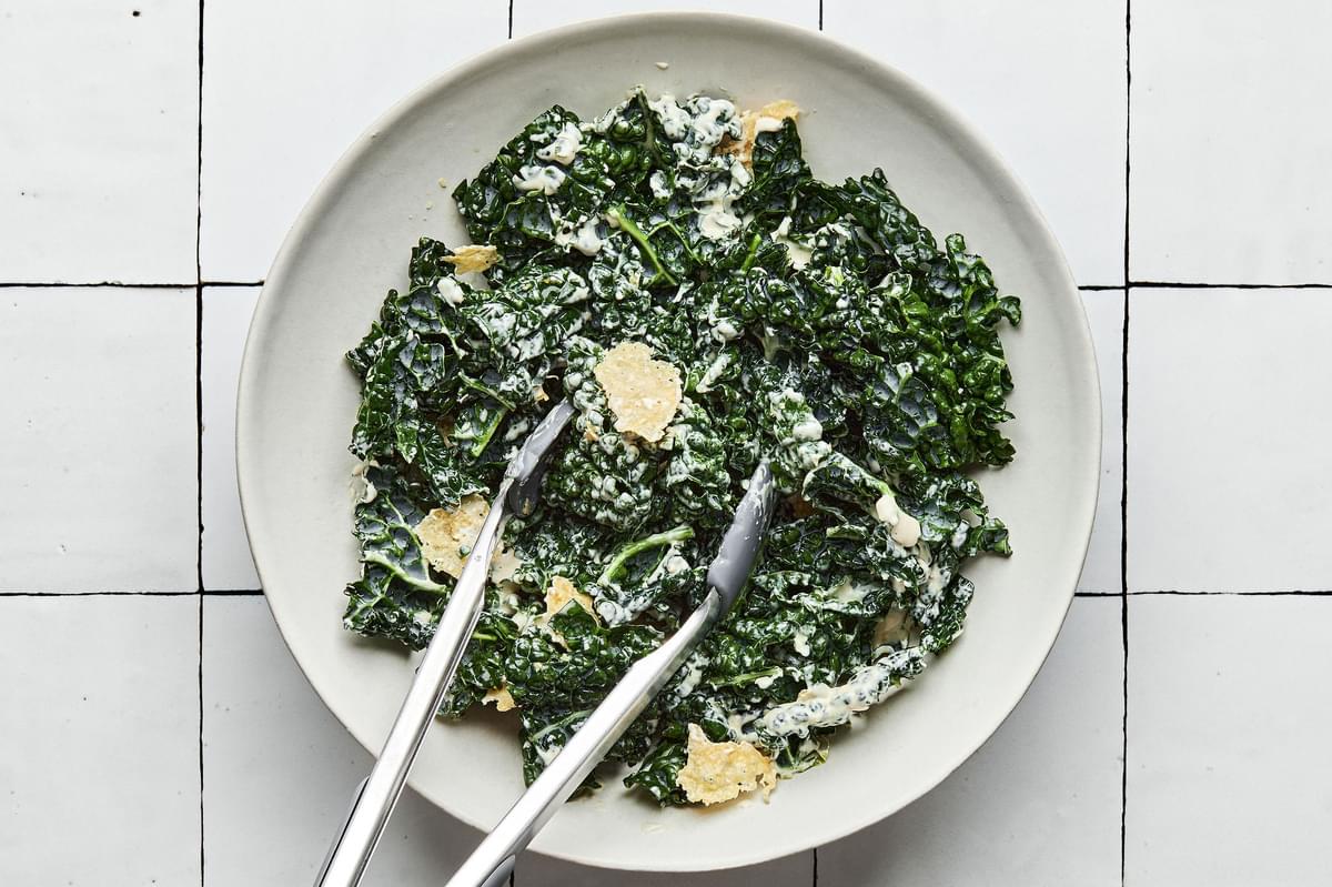 kale, homemade caesar salad, and parmesan crisps tossed in a bowl with tongs