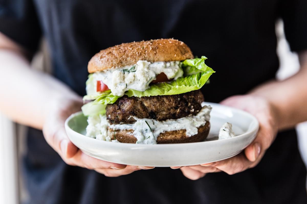Person holding a plate with a Mediterranean Turkey Burger With Tzatziki sauce on it.