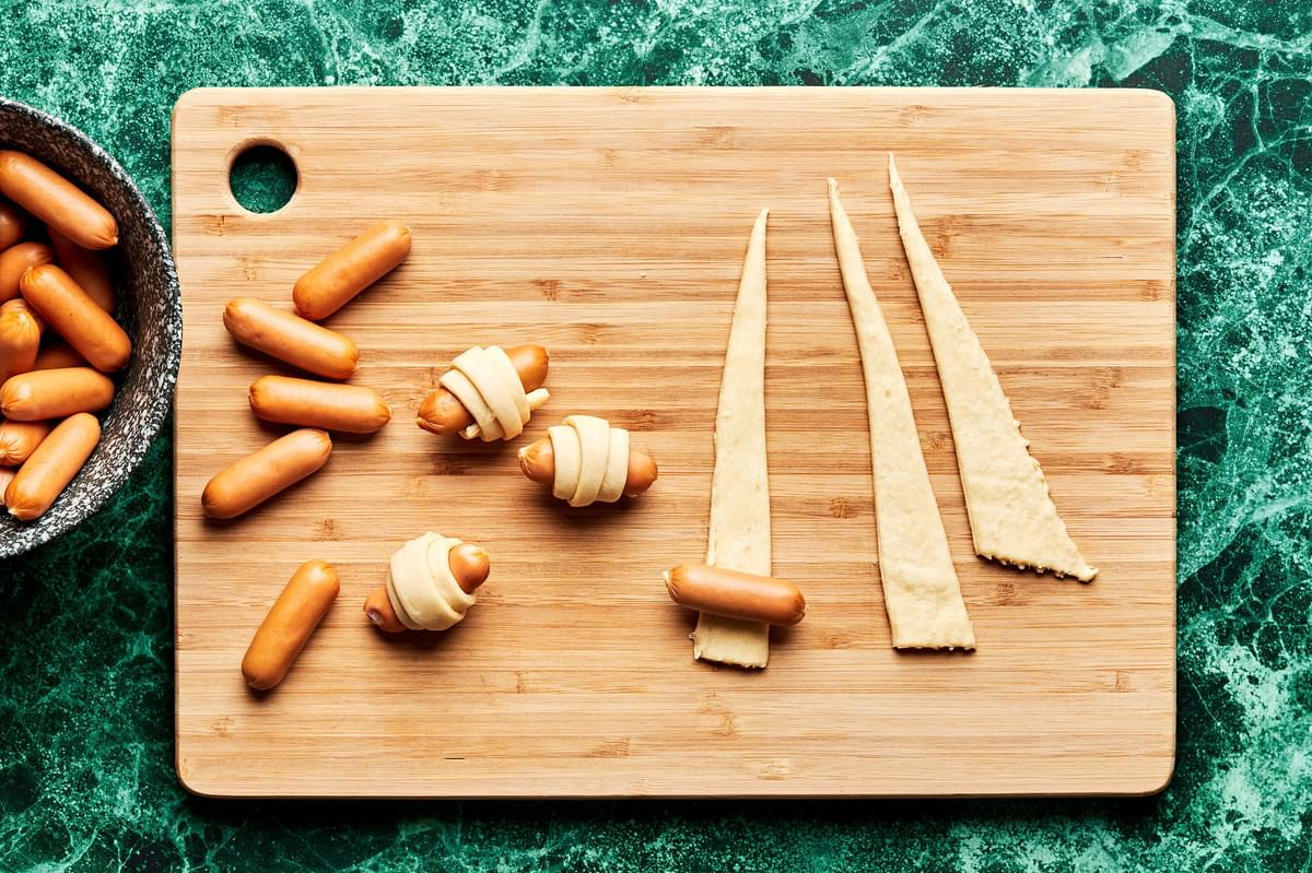 cocktail sausage links being rolled in crescent roll triangles on a cutting board