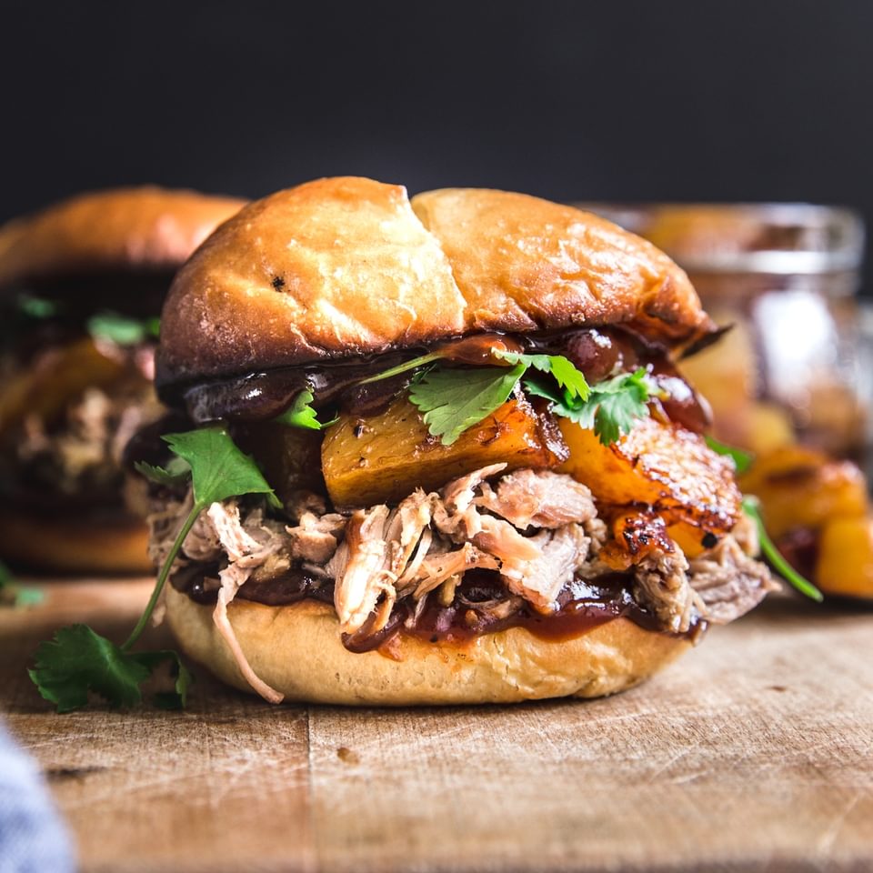 slow cooker pork sandwich with cilantro pineapples, onions and bbq sauce on a bun on a butting board.