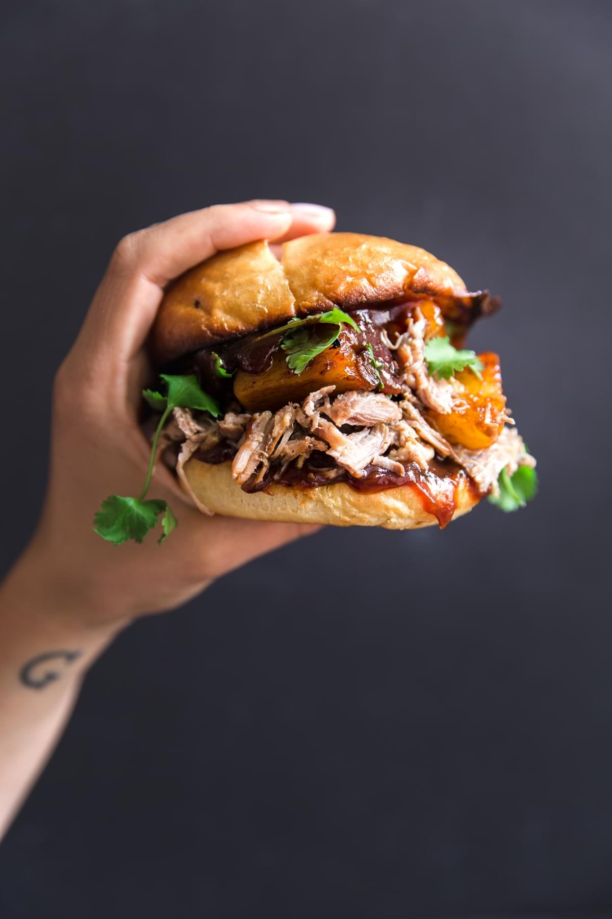 A hand holding a slow cooker pork sandwich with cilantro pineapples, onions and bbq sauce on a bun.