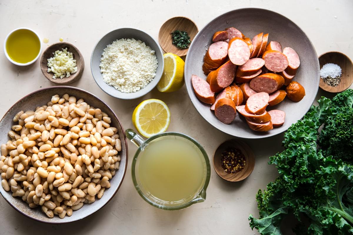 white beans, chicken stock, sausage, kale, parmesan, lemon, thyme, garlic and olive oil on a counter for a skillet dinner