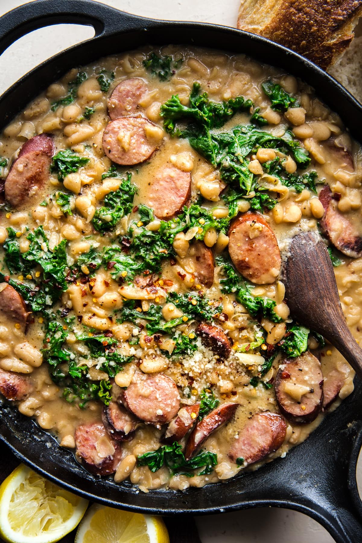 white bean and sausage skillet dinner with kale, parmesan cheese and lemon being stirred with a spoon
