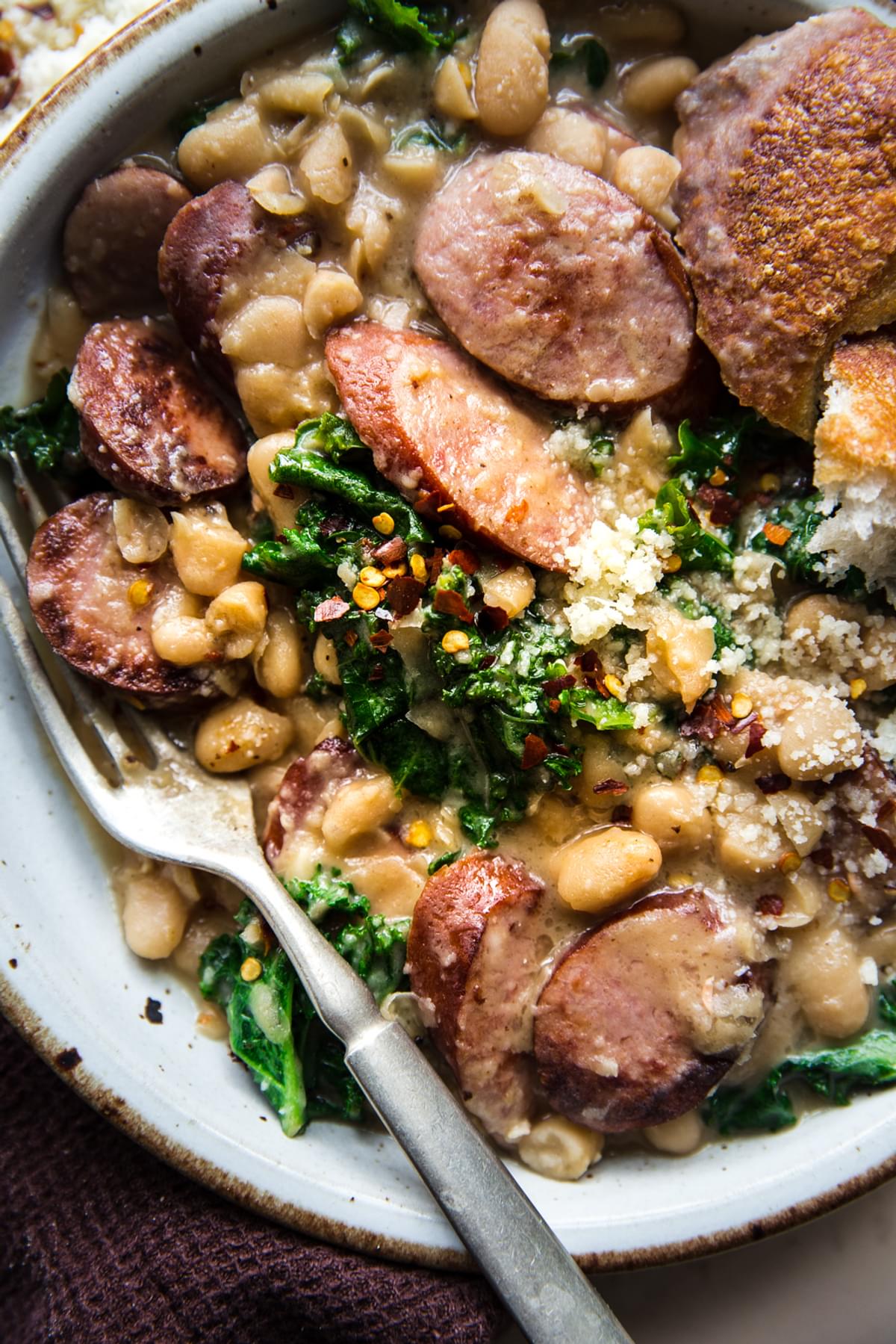 white bean and sausage skillet dinner with kale in a bowl with a fork and red pepper flakes.