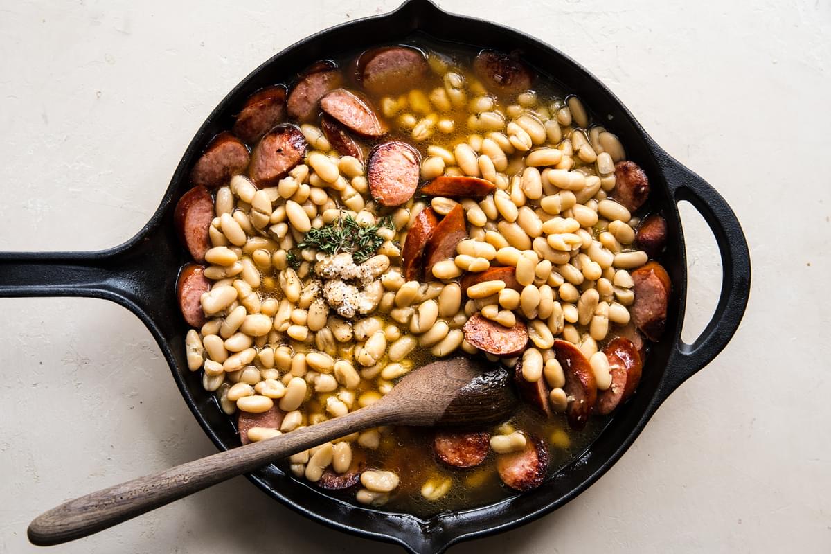 white bean and sausage skillet with chicken stock, garlic and thyme being stored with a wooden spoon.