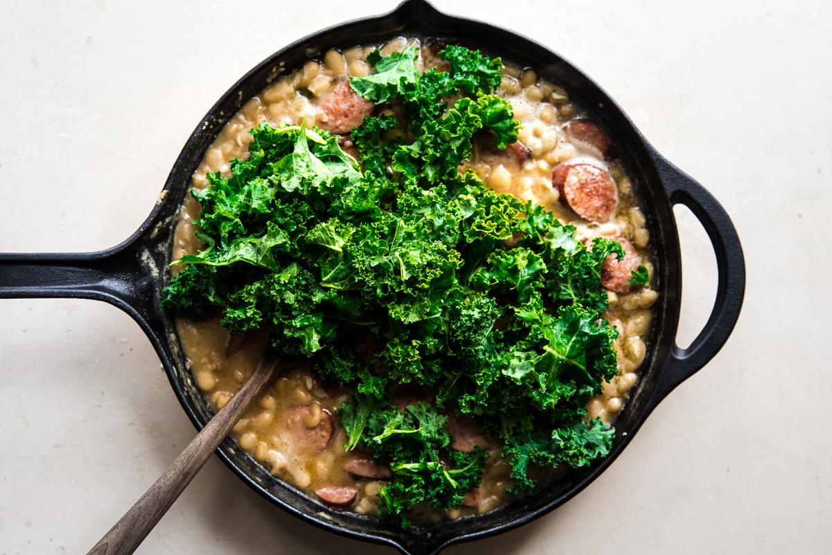 White Bean Sausage dinner with garlic, chicken stock and kale being stirred together with a spoon.