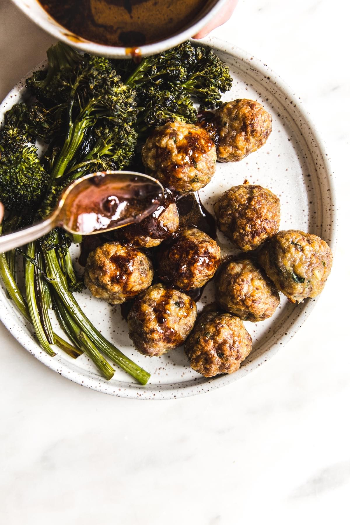 spoon drizzling homemade teriyaki sauce over baked meatballs on a plate with roasted broccoli